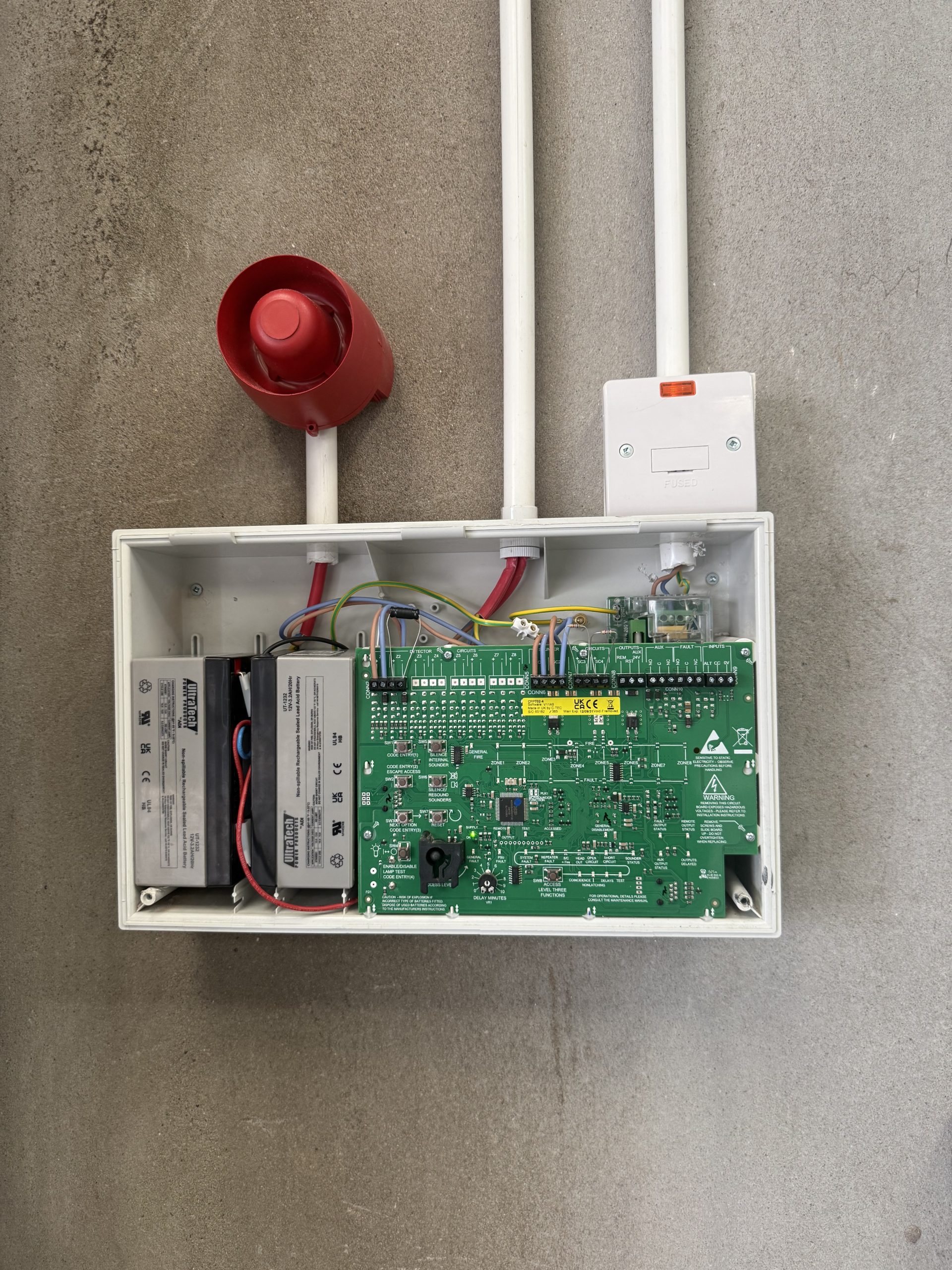 Open fire alarm control panel showing its circuit board and standby batteries, with a red sounder above