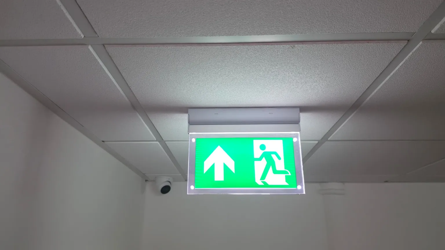 An illuminated green emergency exit sign mounted below a suspended ceiling, with a dome camera on the wall behind