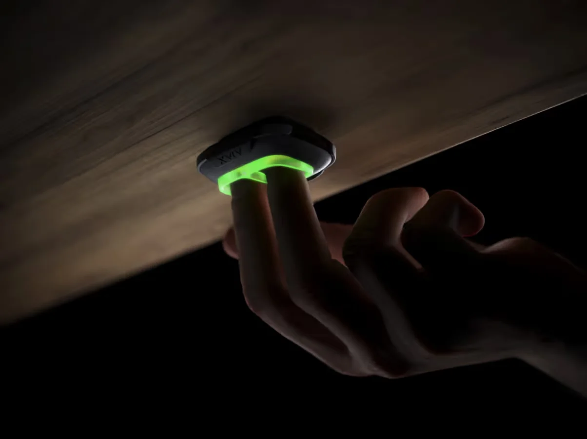 A hand pressing a wireless panic button fixed under a desk, its indicator ring lit