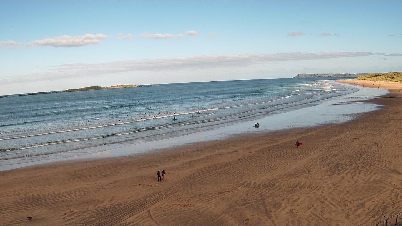 Live view from the Portrush surf camera over East Strand, with surfers in the water and the Skerries on the horizon